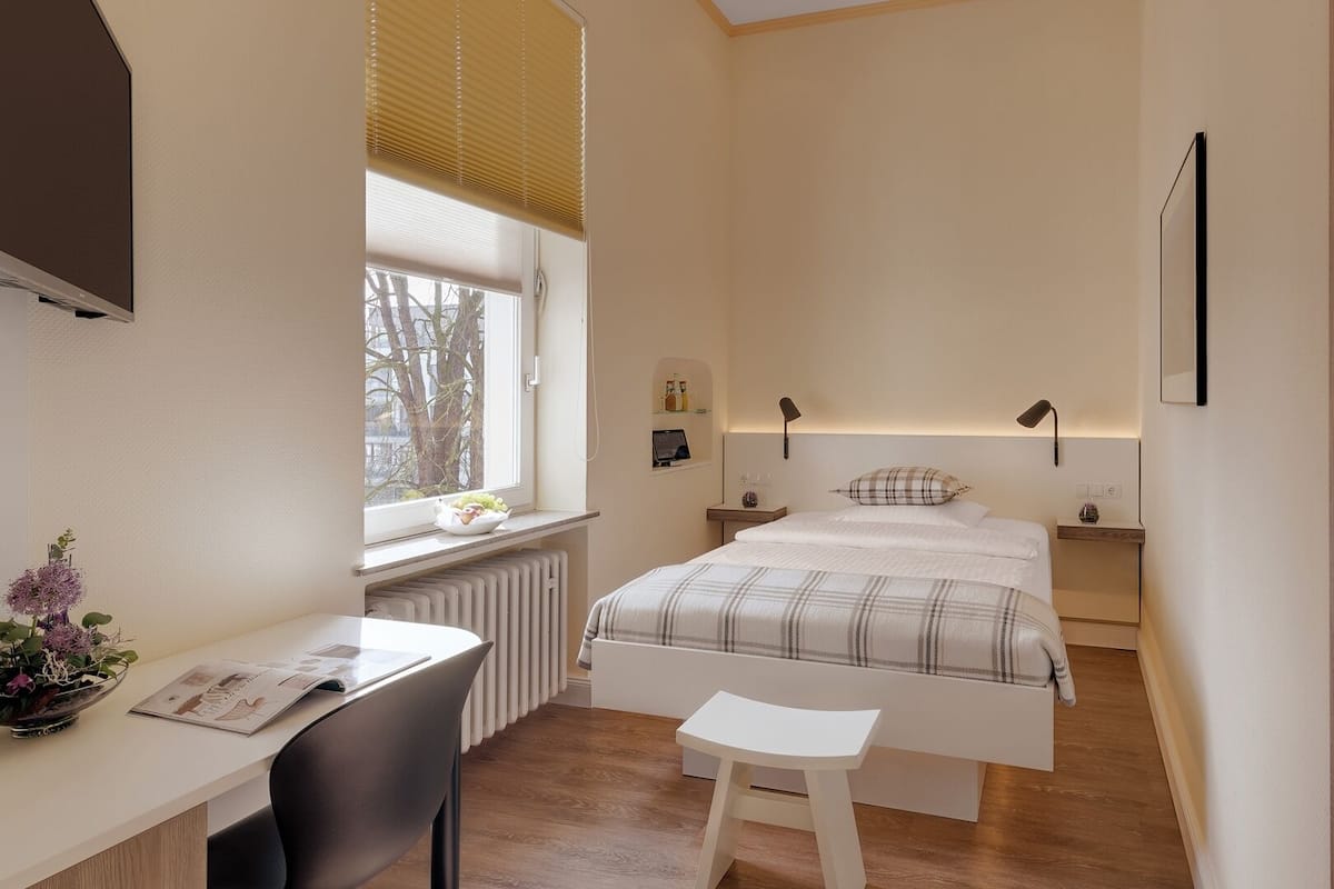A bedroom with a bed, bedside tables, a desk, a chair, a radiator, a window with blinds, and a view of trees outside.