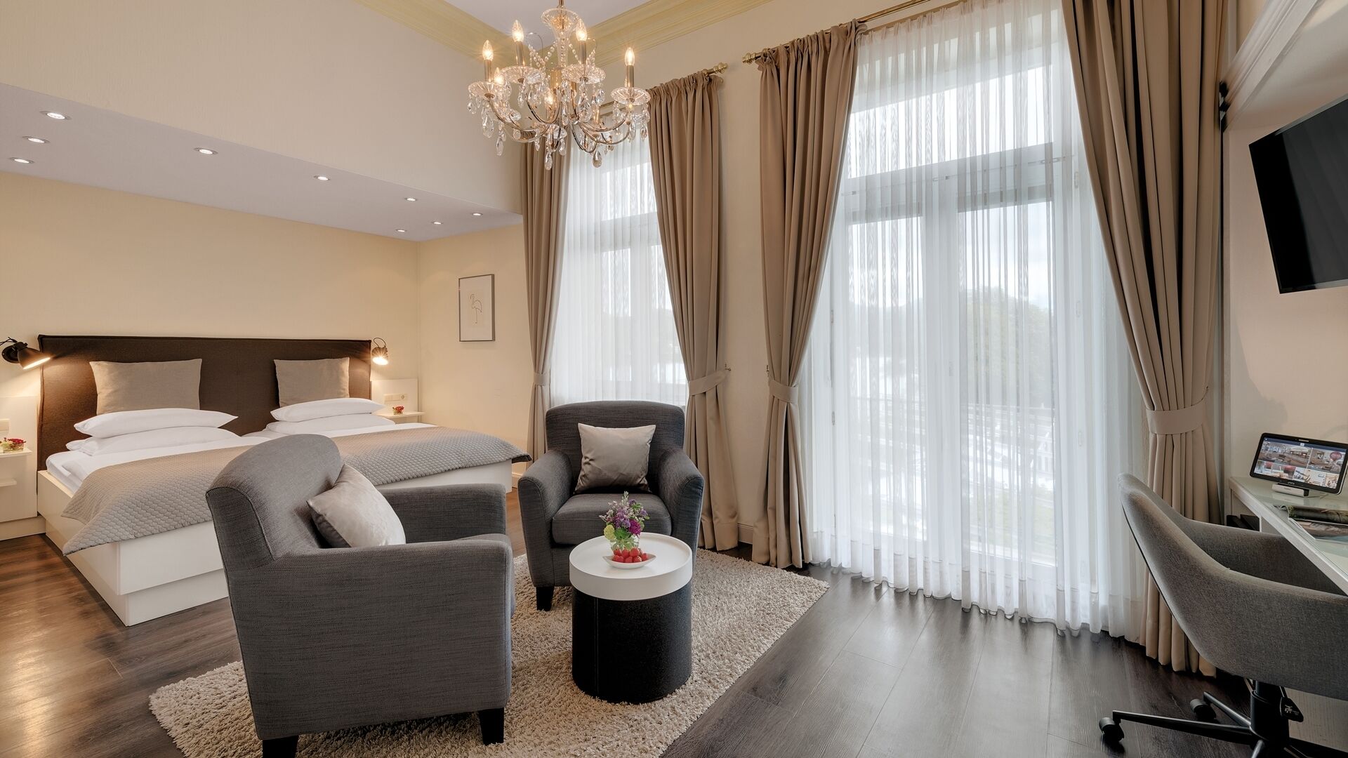Una habitación de hotel moderna con una cama, dos sillones, un escritorio, una araña y ventanas amplias con cortinas.