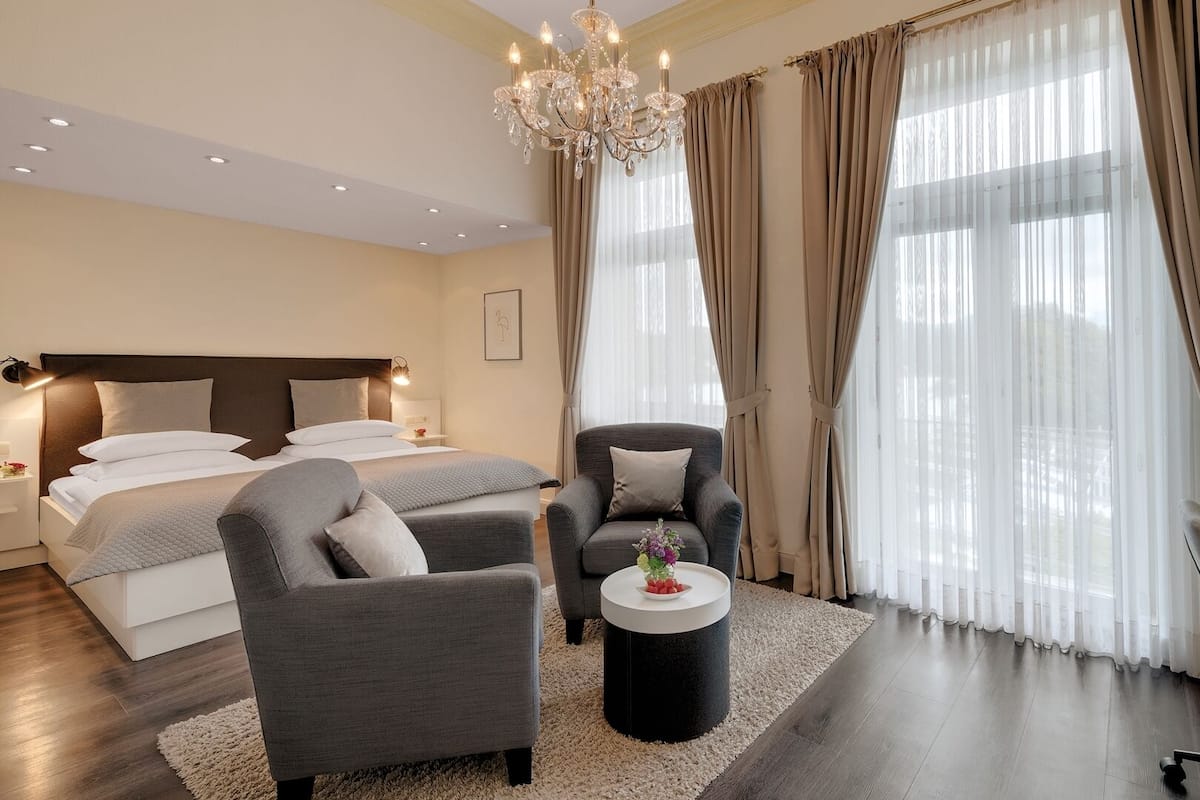 A modern hotel room with a bed, two armchairs, a desk, a chandelier, and large windows with curtains.
