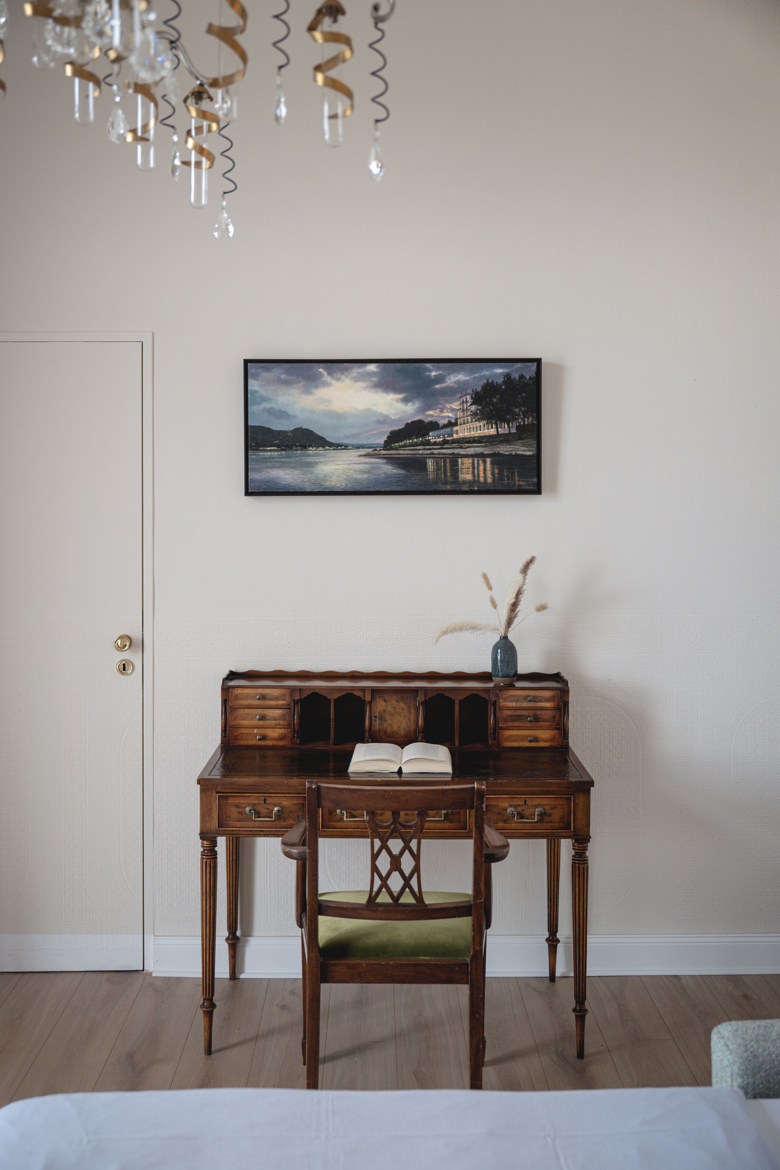 A room with a wooden desk, a chair, a framed landscape painting, and a chandelier.