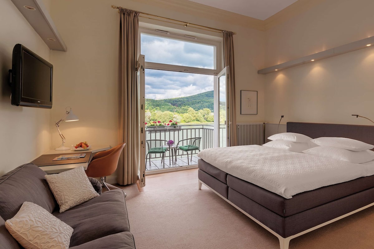 A hotel room with a large bed, a sofa, a desk with a lamp, and a balcony with a view of greenery and a cloudy sky.