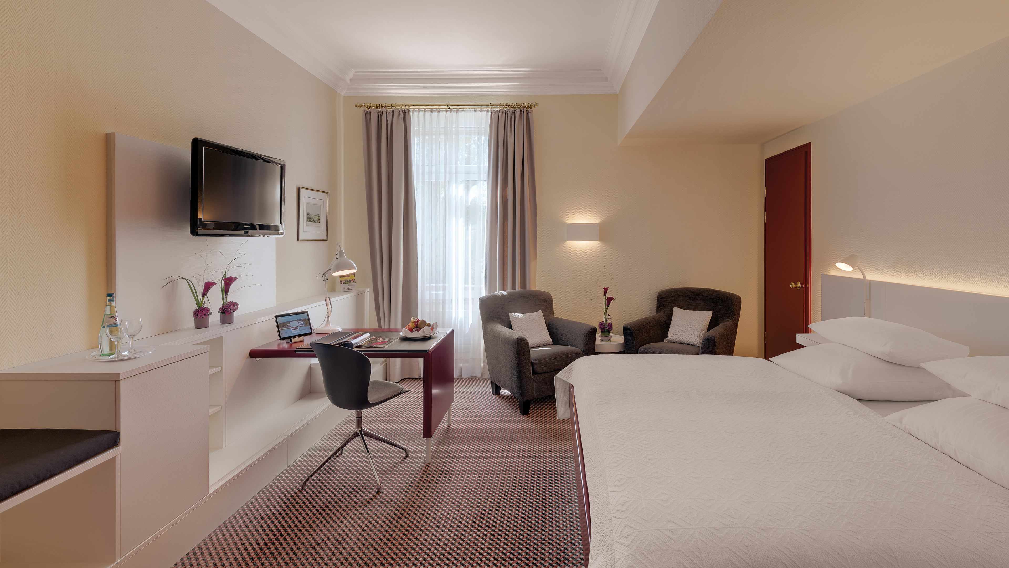 Una habitación de hotel con una cama, dos sillones, un escritorio con una computadora, un televisor montado en la pared y una ventana con cortinas.