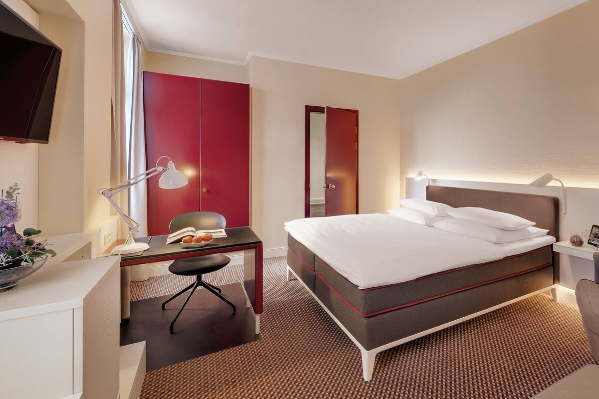A hotel room with a bed, desk, chair, lamp, and a TV.