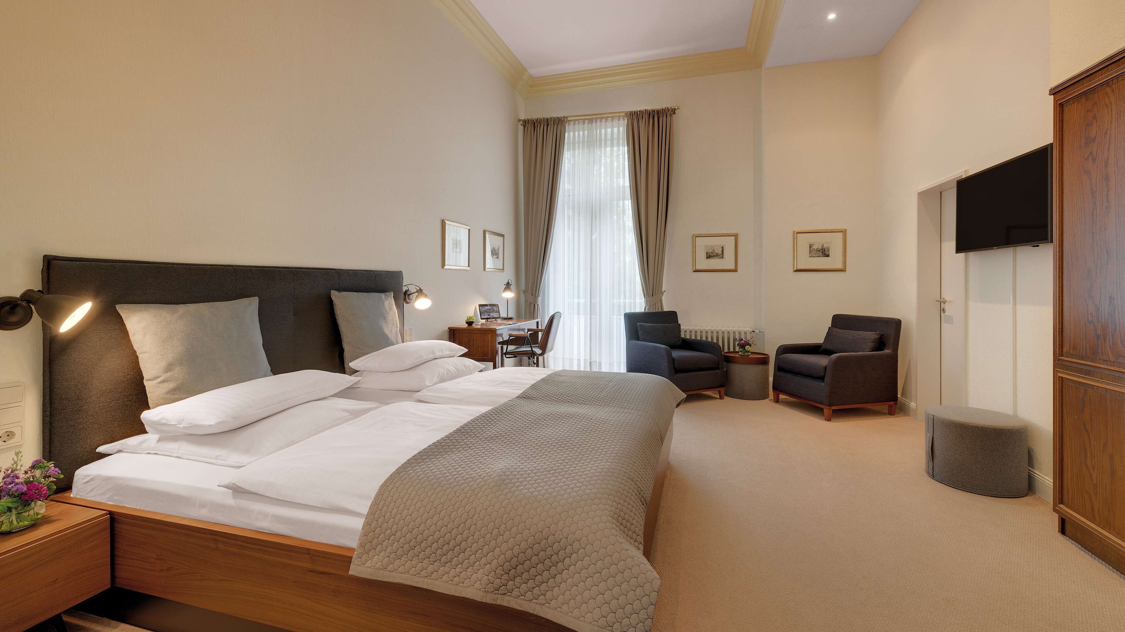 Habitación de hotel con una cama grande, dos sillones, un escritorio con una silla, un televisor y cuadros en la pared.