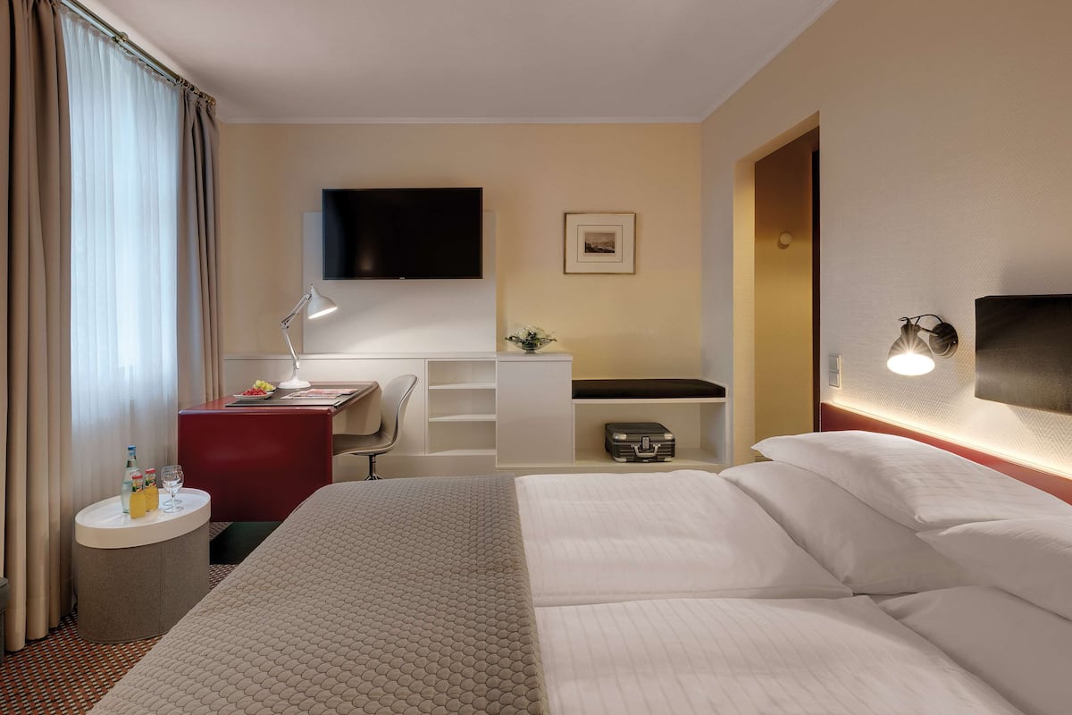 A hotel room with a large bed, a desk with a chair, a TV mounted on the wall, a bedside table with a lamp, and a small shelf with a suitcase.