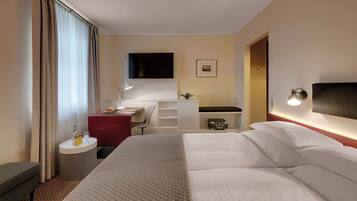 A hotel room with a large bed, a desk with a chair, a TV mounted on the wall, a bedside table with a lamp, and a small shelf with a suitcase.