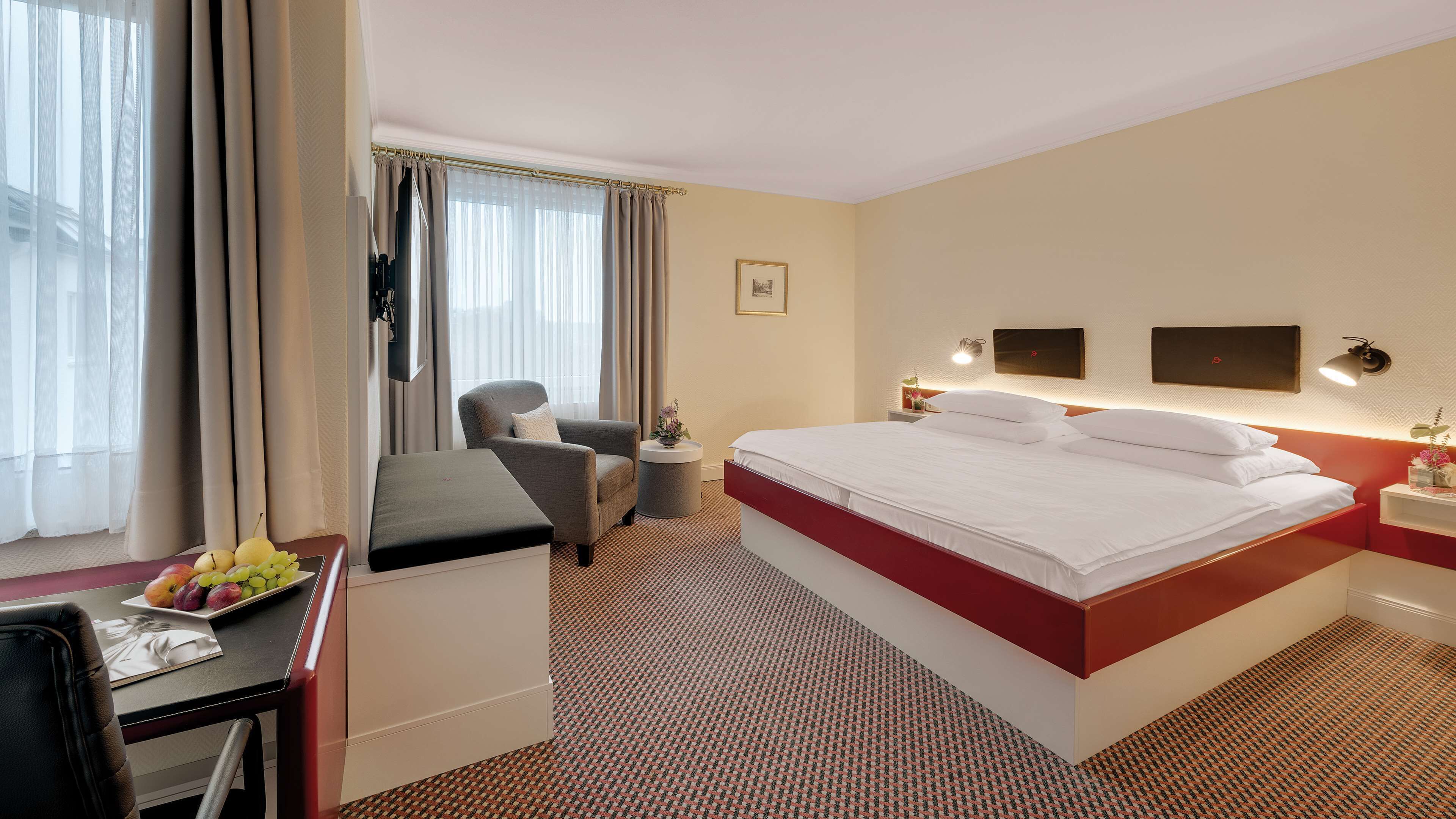 A hotel room with a bed, a desk with a chair, a small table with a vase, and a window with curtains.