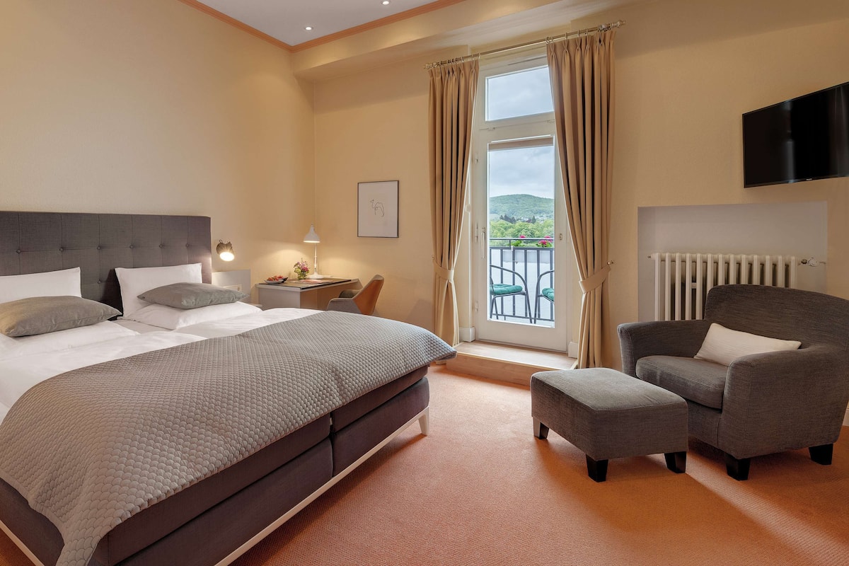 A bedroom with a large bed, two bedside tables, a chair, a desk, a radiator, and a TV.