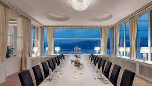 Una larga mesa de comedor dispuesta para una comida formal, con sillas, una araña y ventanas amplias con cortinas.