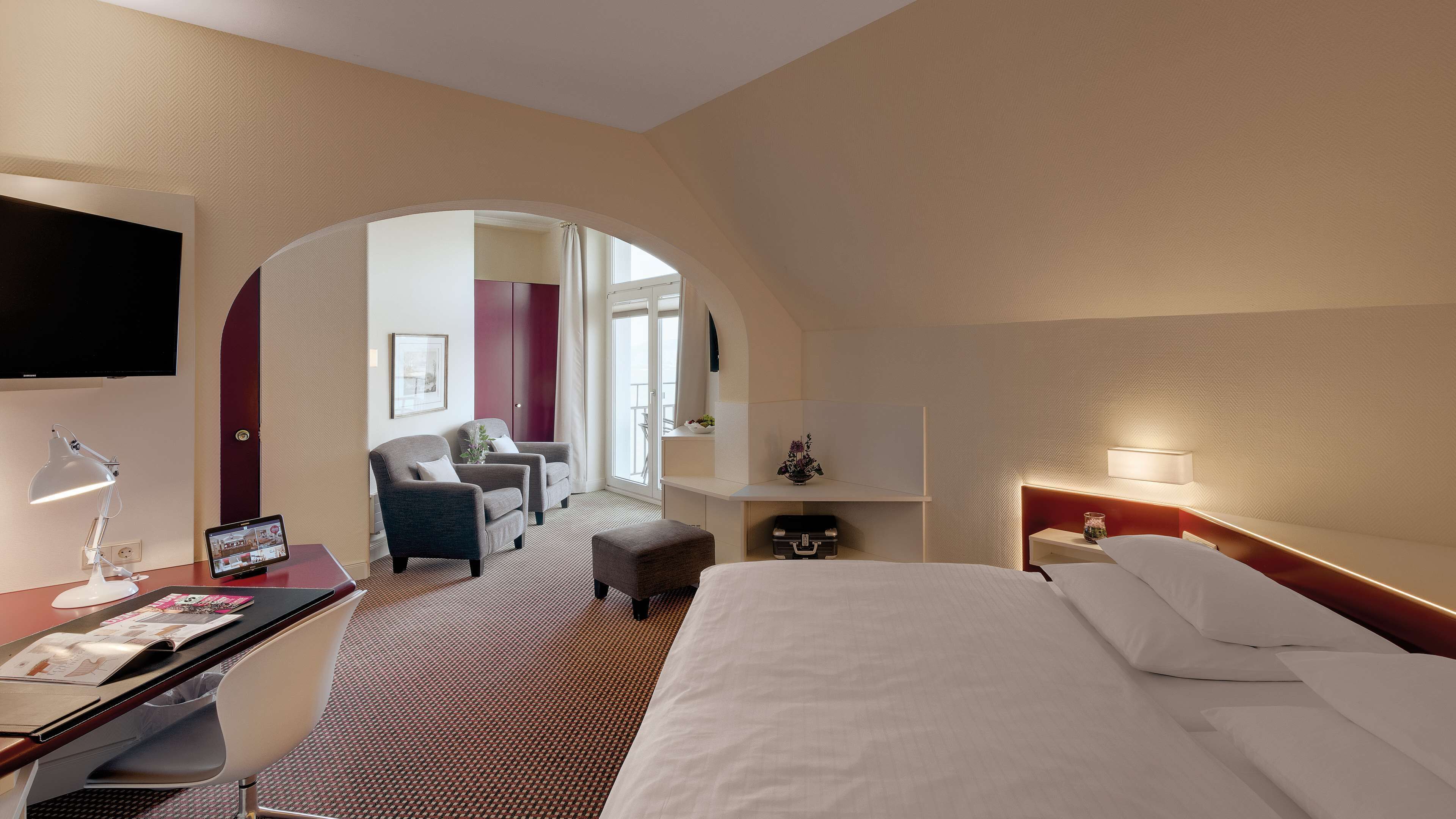 Hotelkamer met een bed, bureau, stoel, televisie en uitzicht door een boogvormige deuropening naar een balkon.