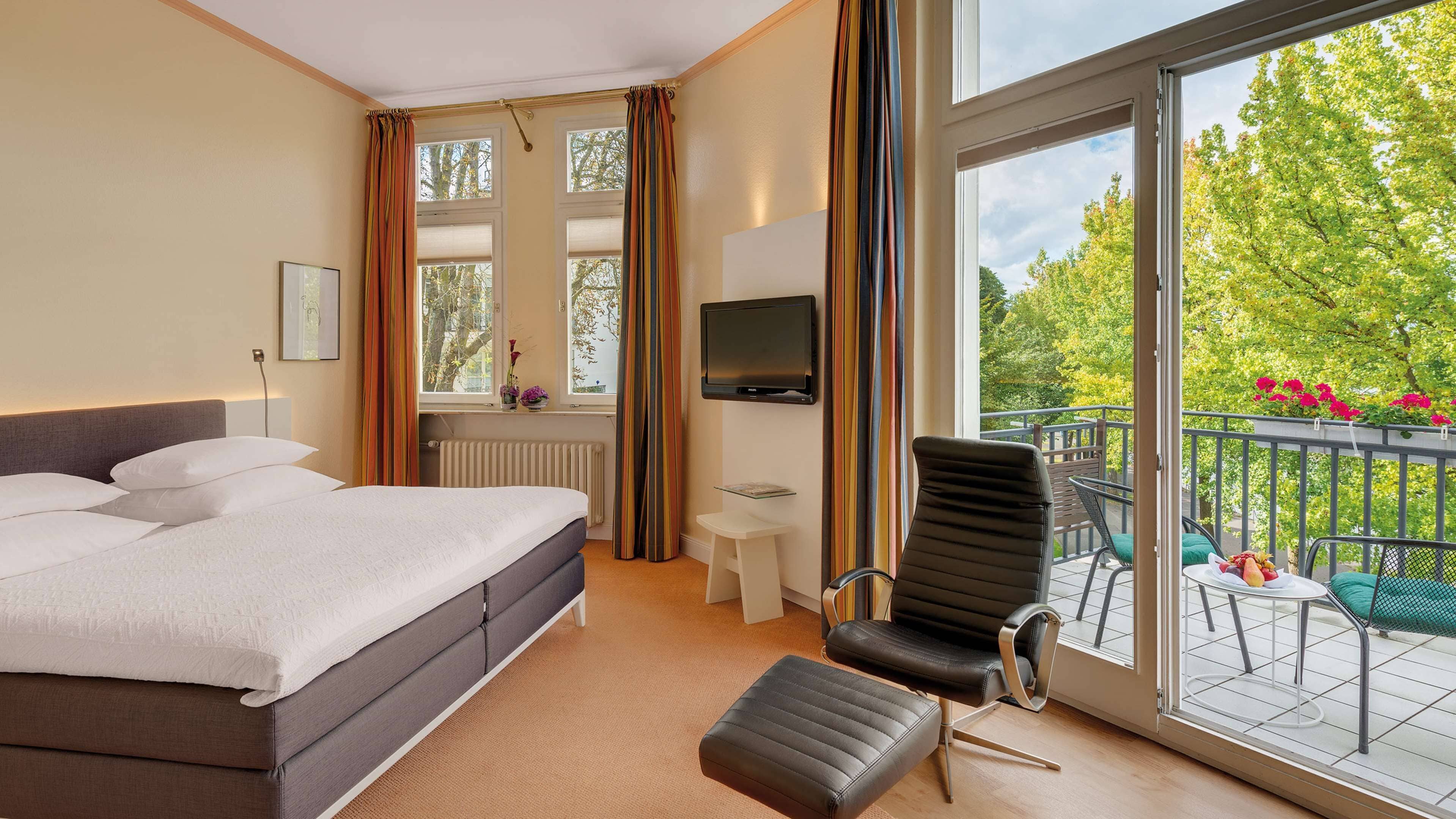 Ein Hotelzimmer mit Bett, Fernseher, Sessel, kleinem Tisch und einem Balkon mit Blick auf Bäume und Blumen.