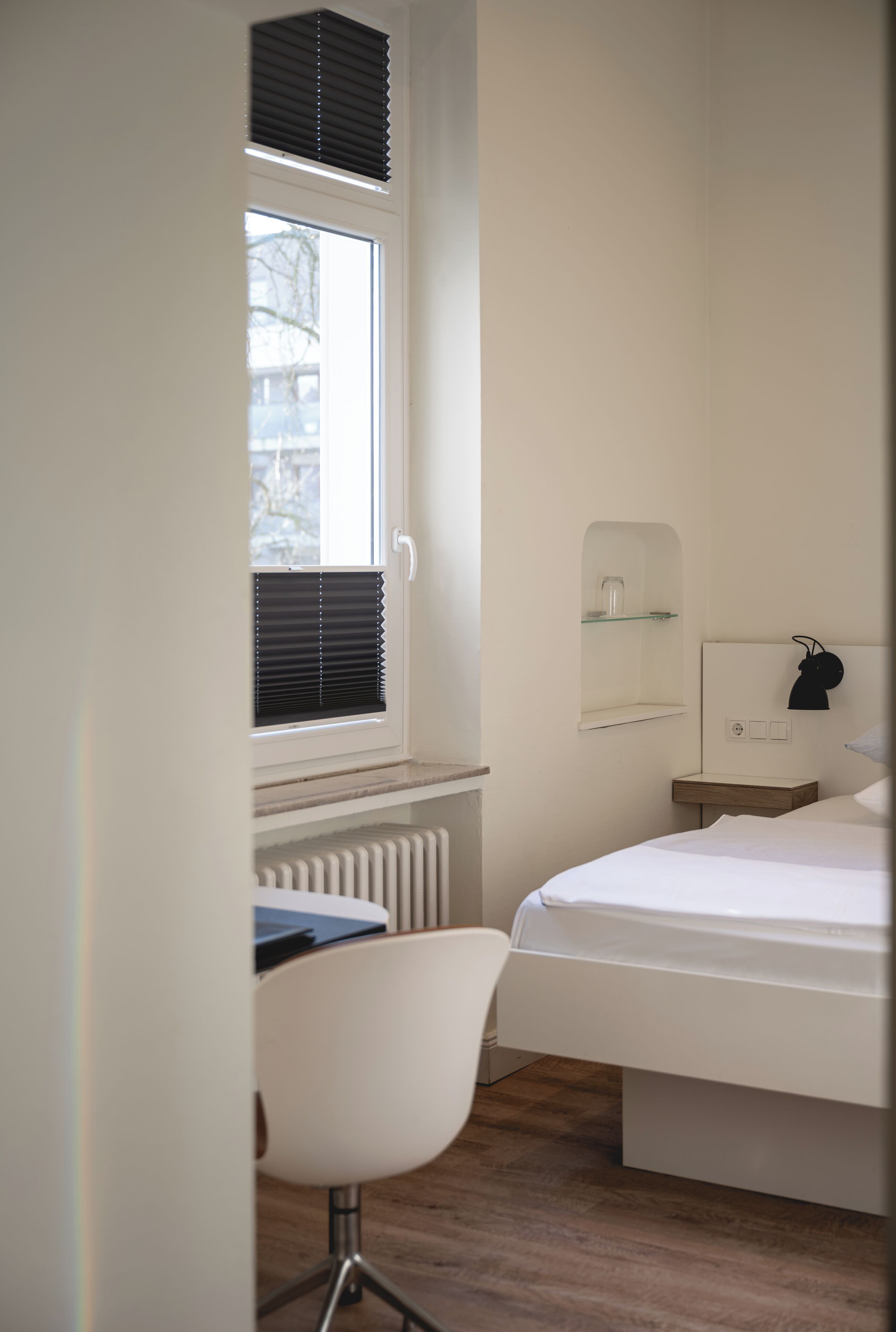 A bedroom with a bed, a chair, a window with blinds, a radiator, and a shelf with a lamp.