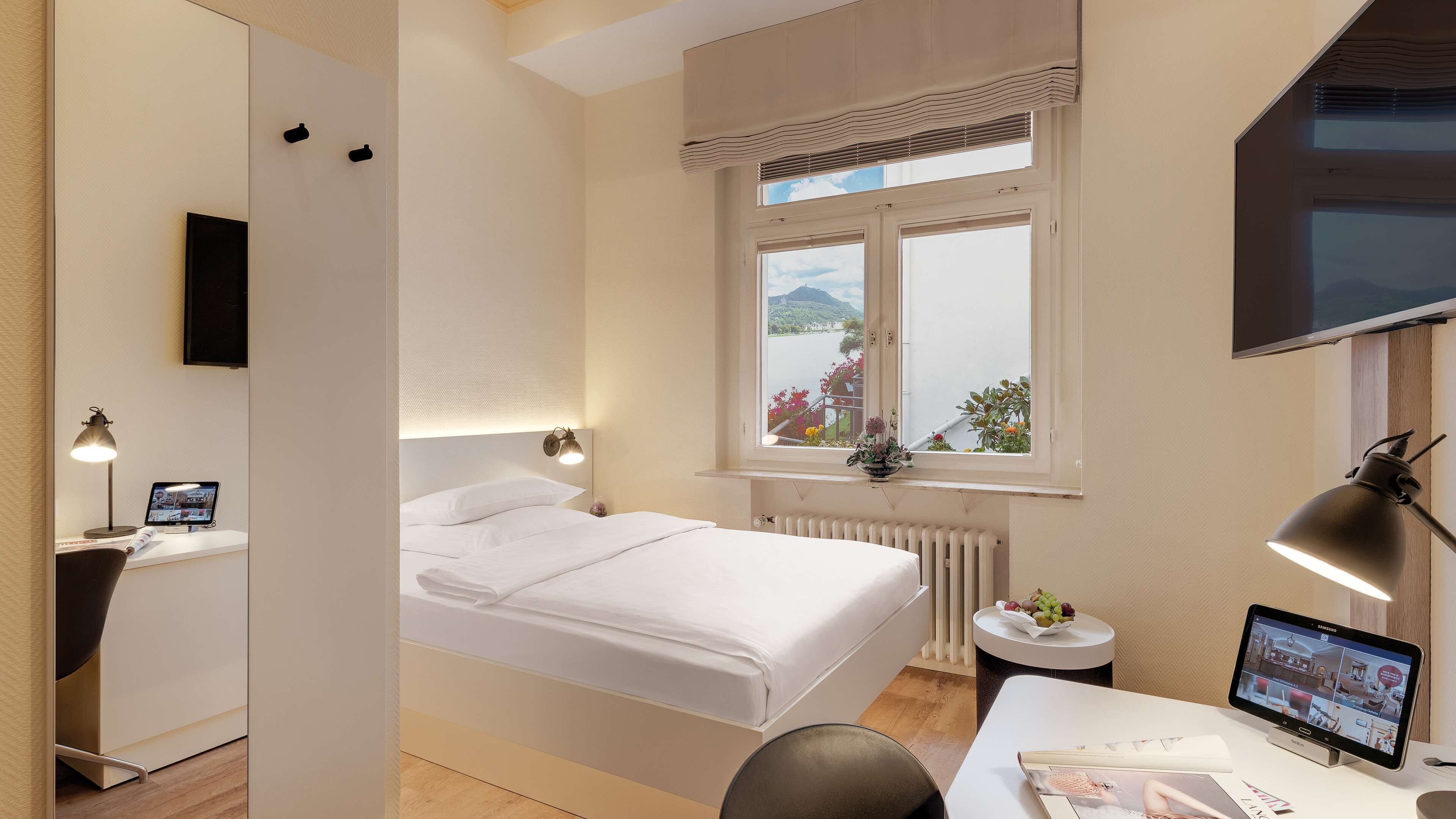 Ein modernes Hotelzimmer mit einem Bett, einem Schreibtisch mit Computer, einem kleinen Tisch mit Obstschale und einem Fenster mit Blick ins Freie.