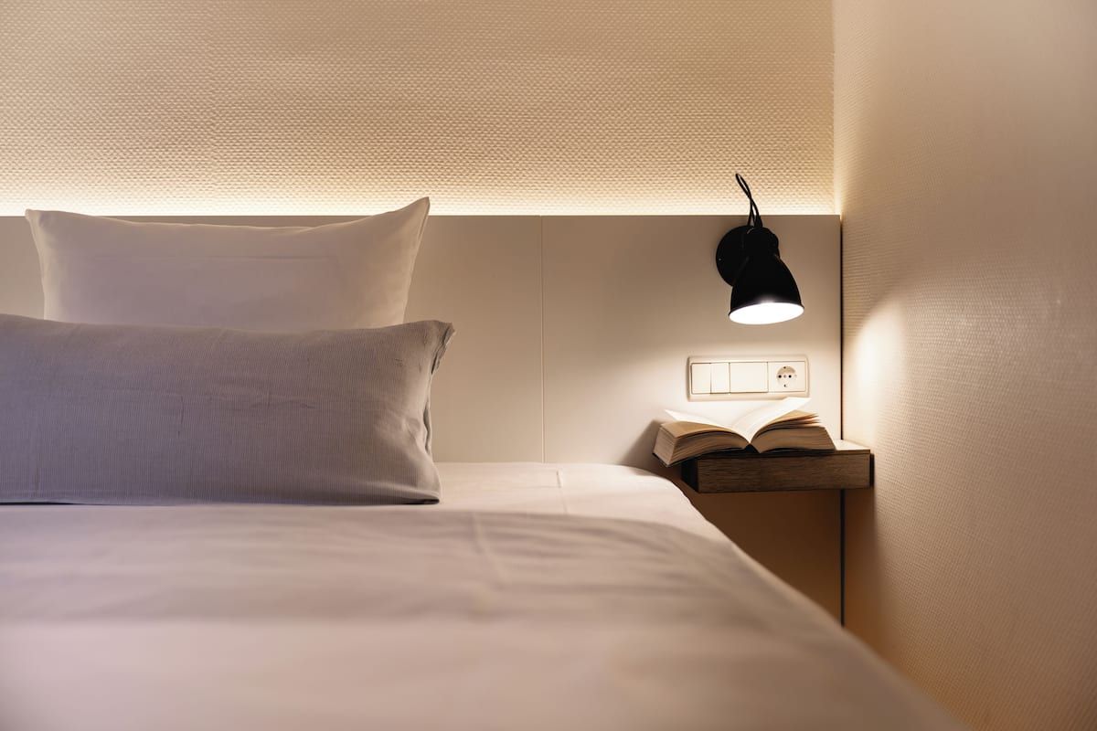 A neatly made bed with pillows, a bedside table with a book and a lamp, and a wall socket.