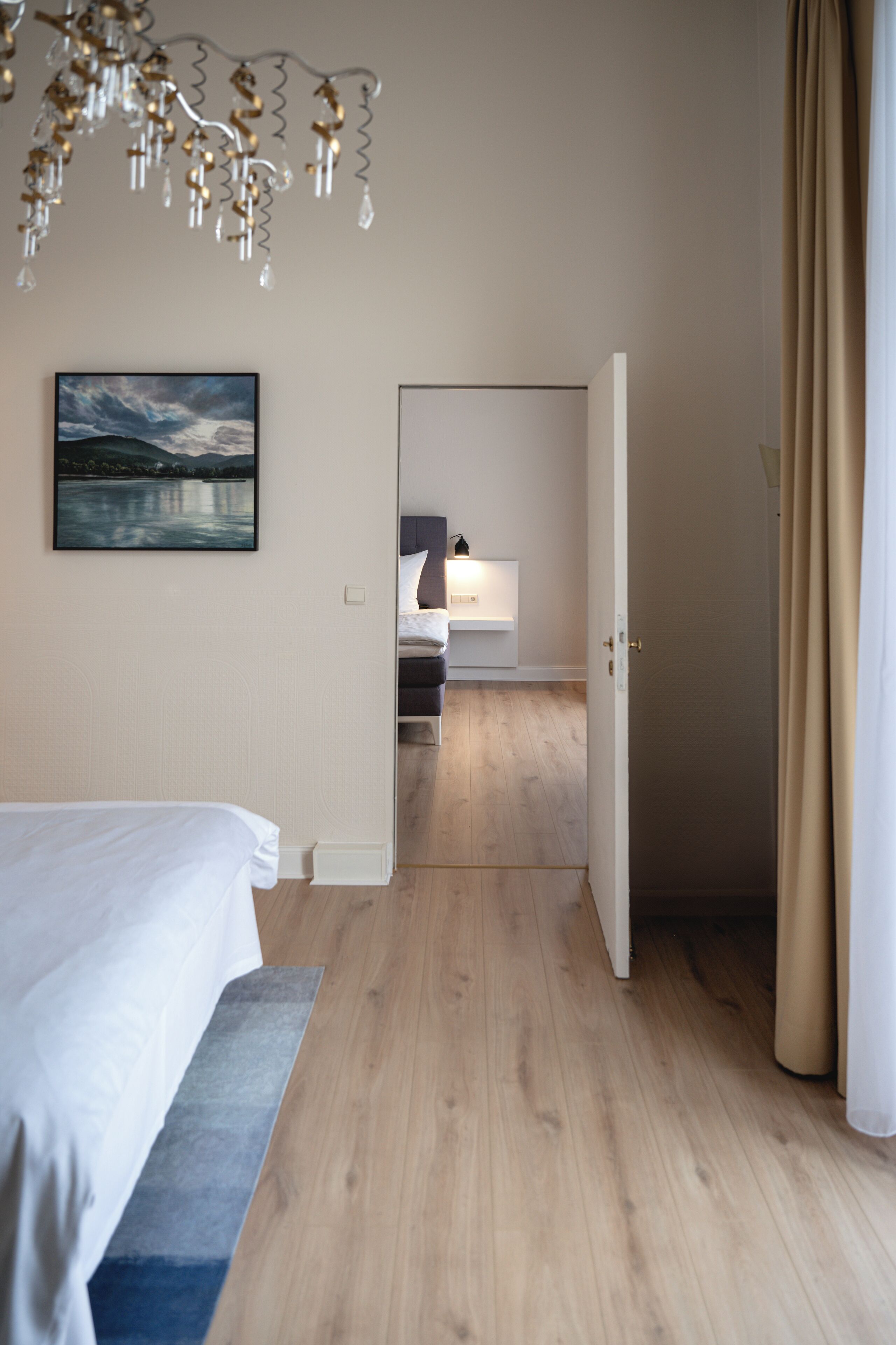 Habitació d'hotel amb un llit, un quadre, un sòl de fusta i un bany visible a través d'una porta oberta.