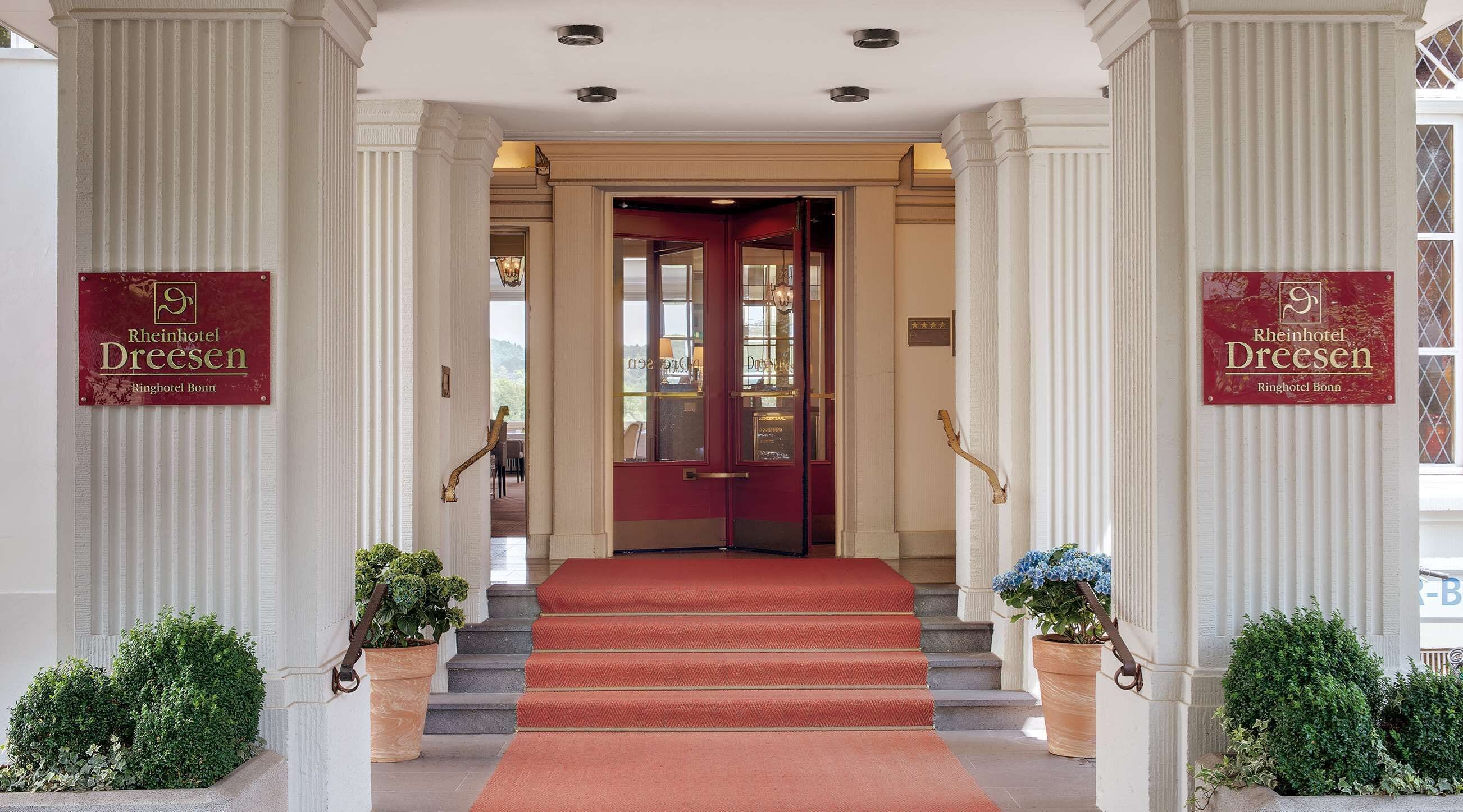 A grand entrance with a red carpet leading to a double door, flanked by columns and potted plants.