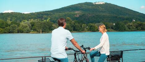 Dos personas junto a un lago, con bicicletas, una baranda metálica y una montaña al fondo.