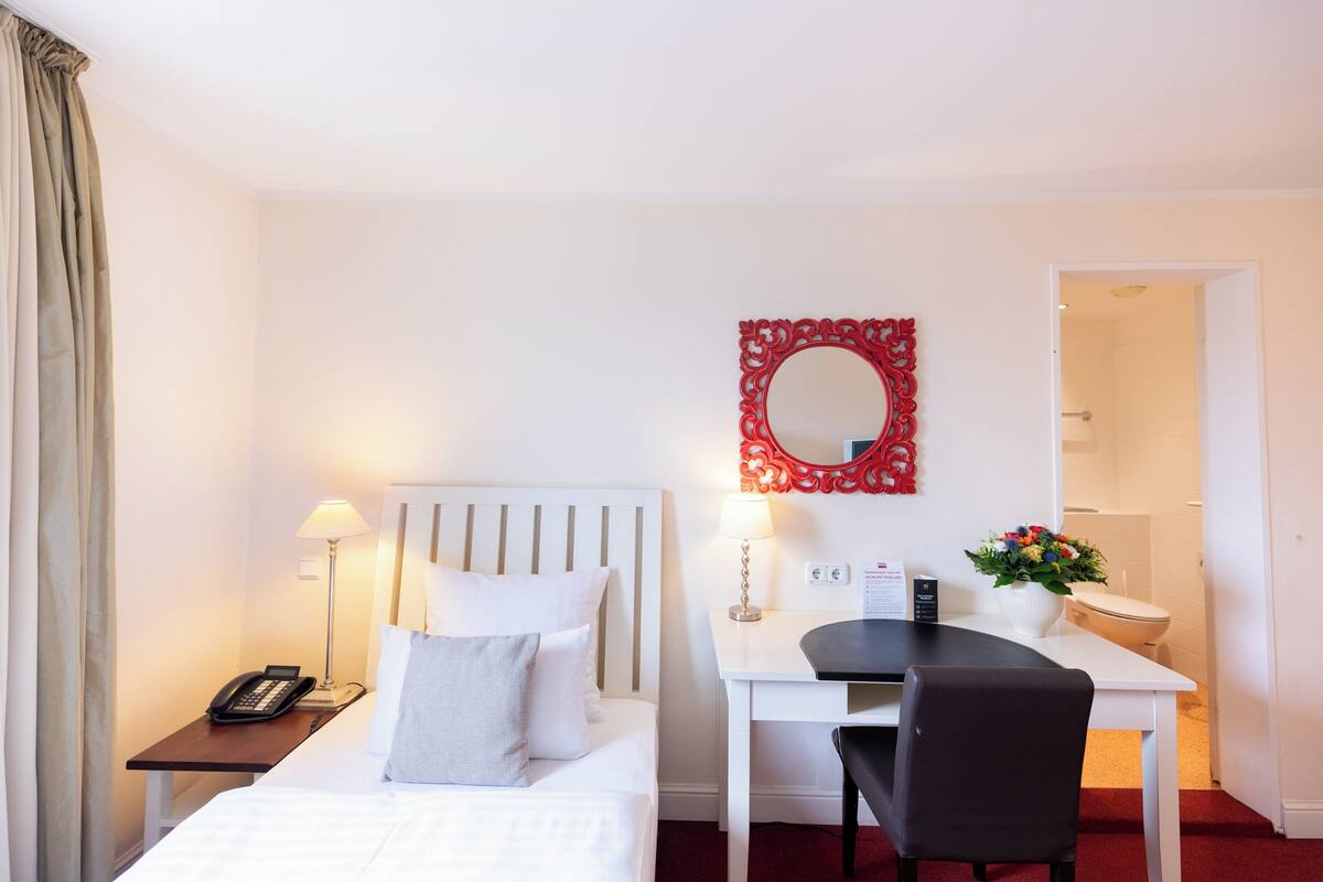 A hotel room with a bed, bedside table with a phone, a desk with a chair, a mirror, and a bathroom visible through an open door.