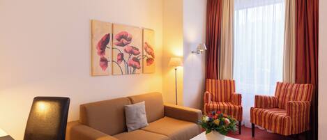 A hotel room with a beige sofa, striped armchairs, a white coffee table with flowers, a lamp, and a painting of flowers on the wall.