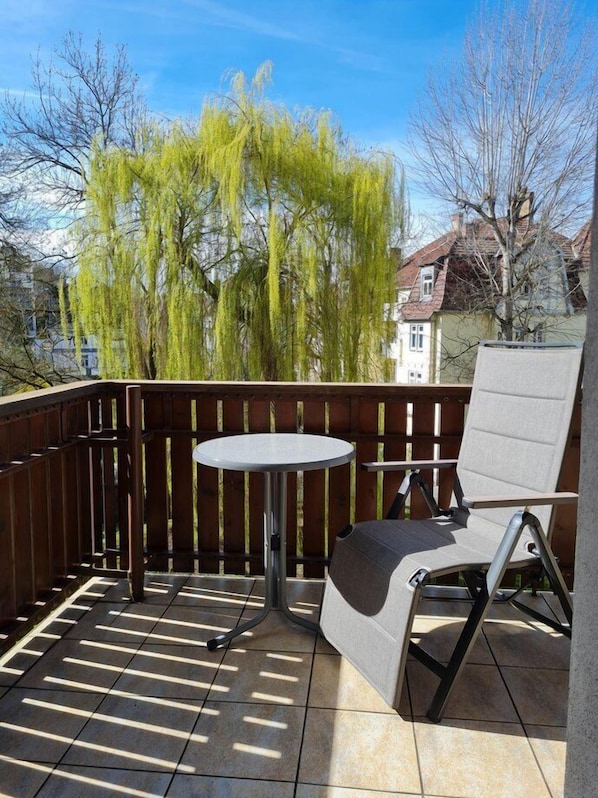 Un balcon avec une table ronde et une seule chaise, entouré d’une clôture en bois, d’où l’on peut admirer des arbres et des bâtiments en arrière-plan.