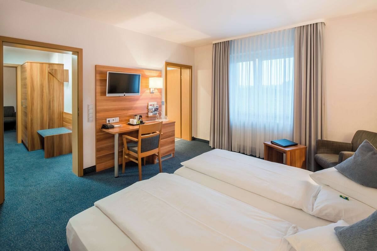 Une chambre d’hôtel avec deux lits, un bureau avec une chaise, un téléviseur fixé au mur et une grande fenêtre avec des rideaux.