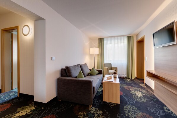 A hotel room with a sofa, a small table, a TV, and a window with curtains. - Best Western Blankenburg Hotel (Coburg)