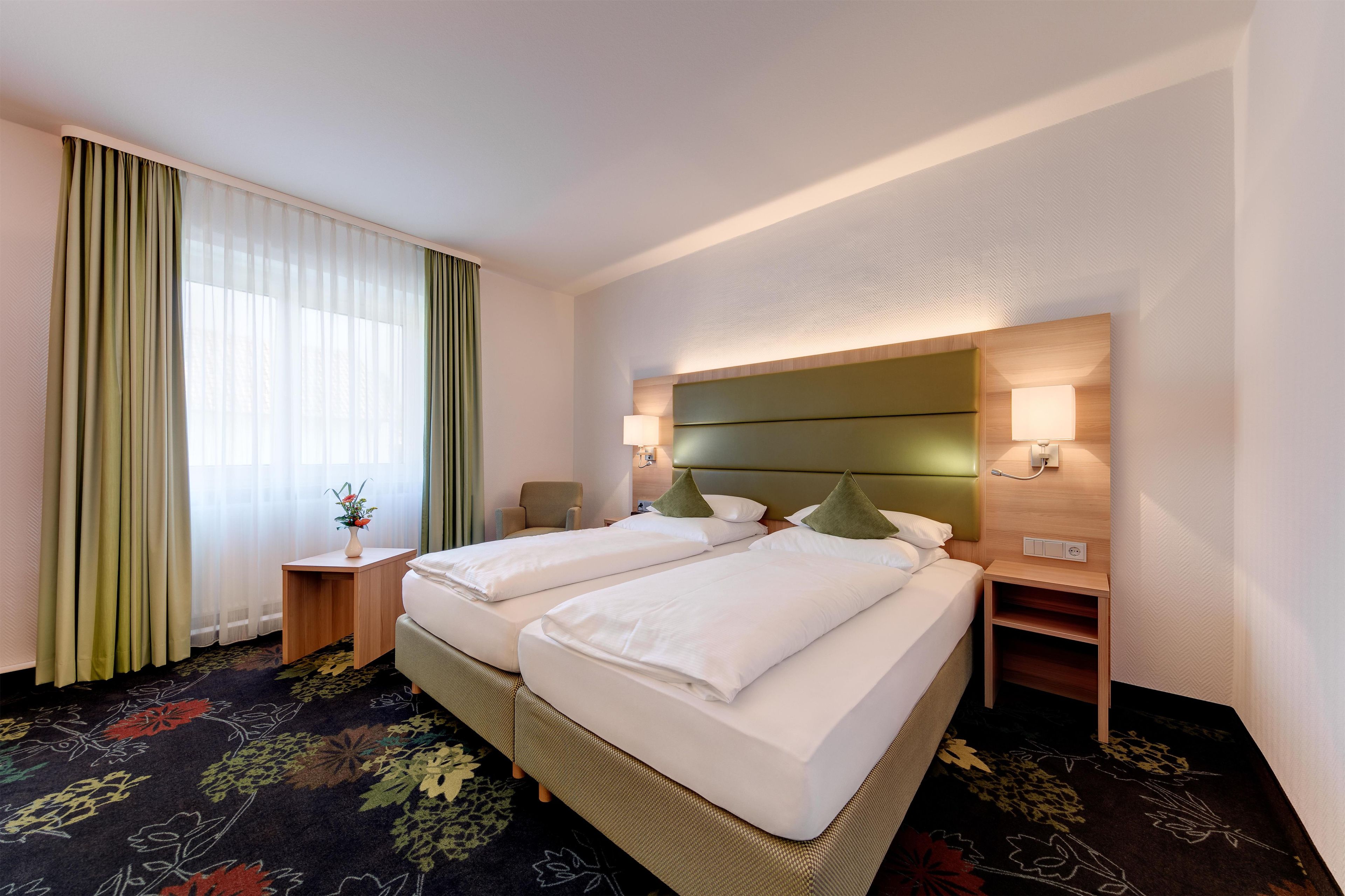 A hotel room with two beds, a nightstand, a lamp, and a window with curtains.