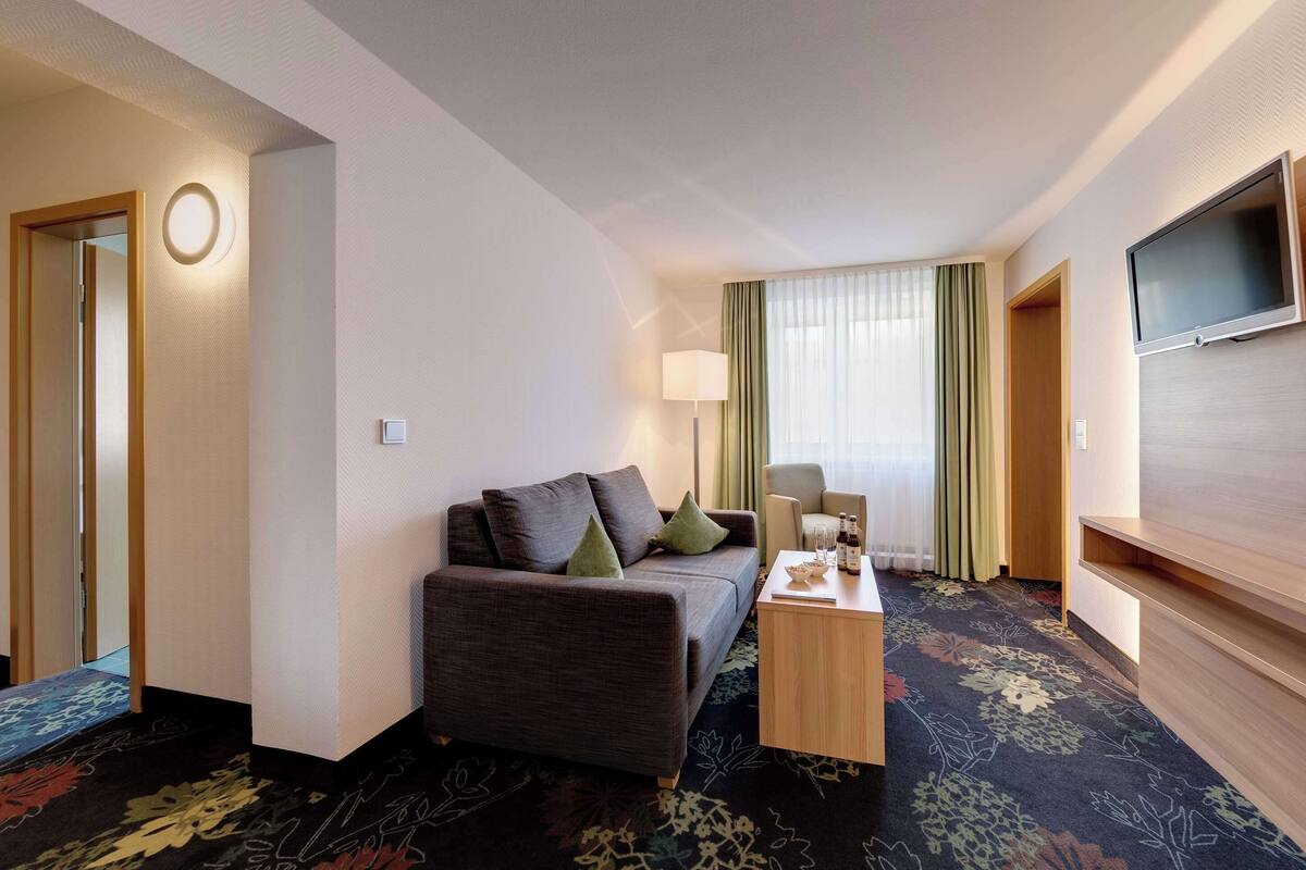 Une chambre d’hôtel avec un canapé, une petite table sur laquelle se trouvent une bouteille et des verres, un téléviseur fixé au mur et une fenêtre avec des rideaux.