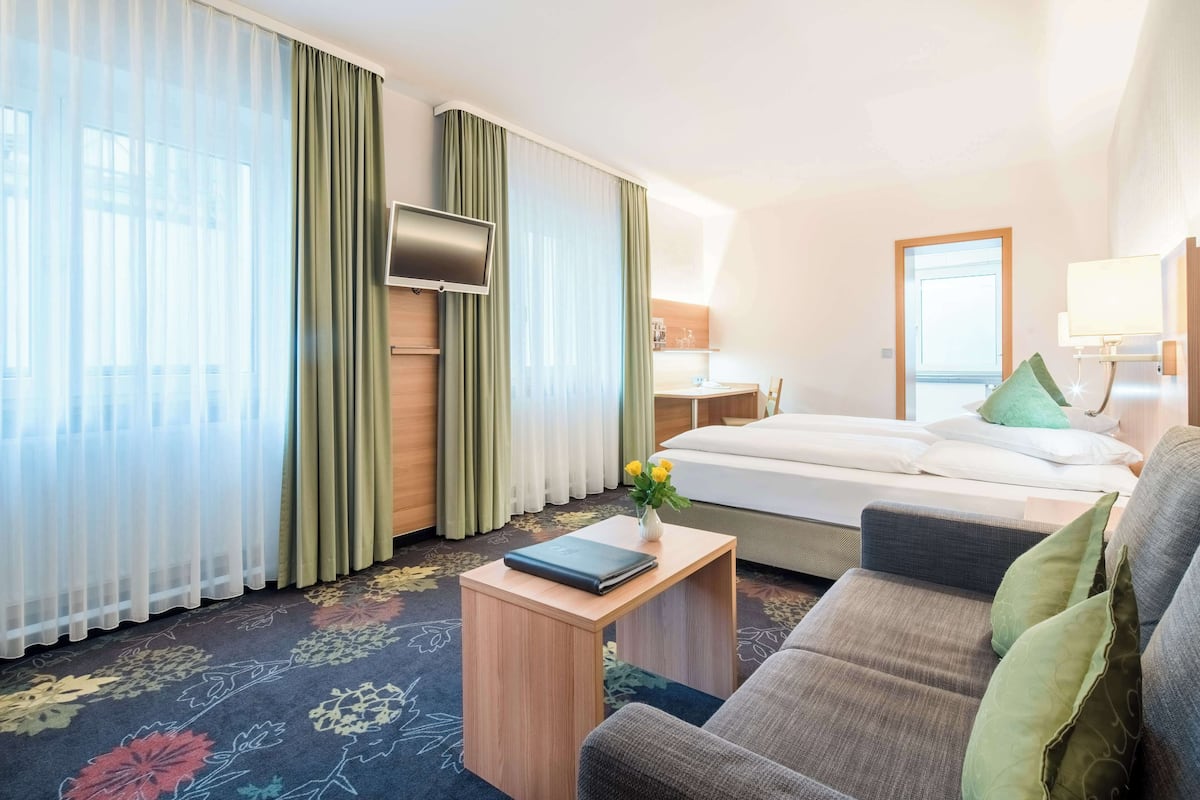 Une chambre d’hôtel avec un lit, un canapé, une petite table avec un livre, une télévision et une fenêtre avec des rideaux légers.