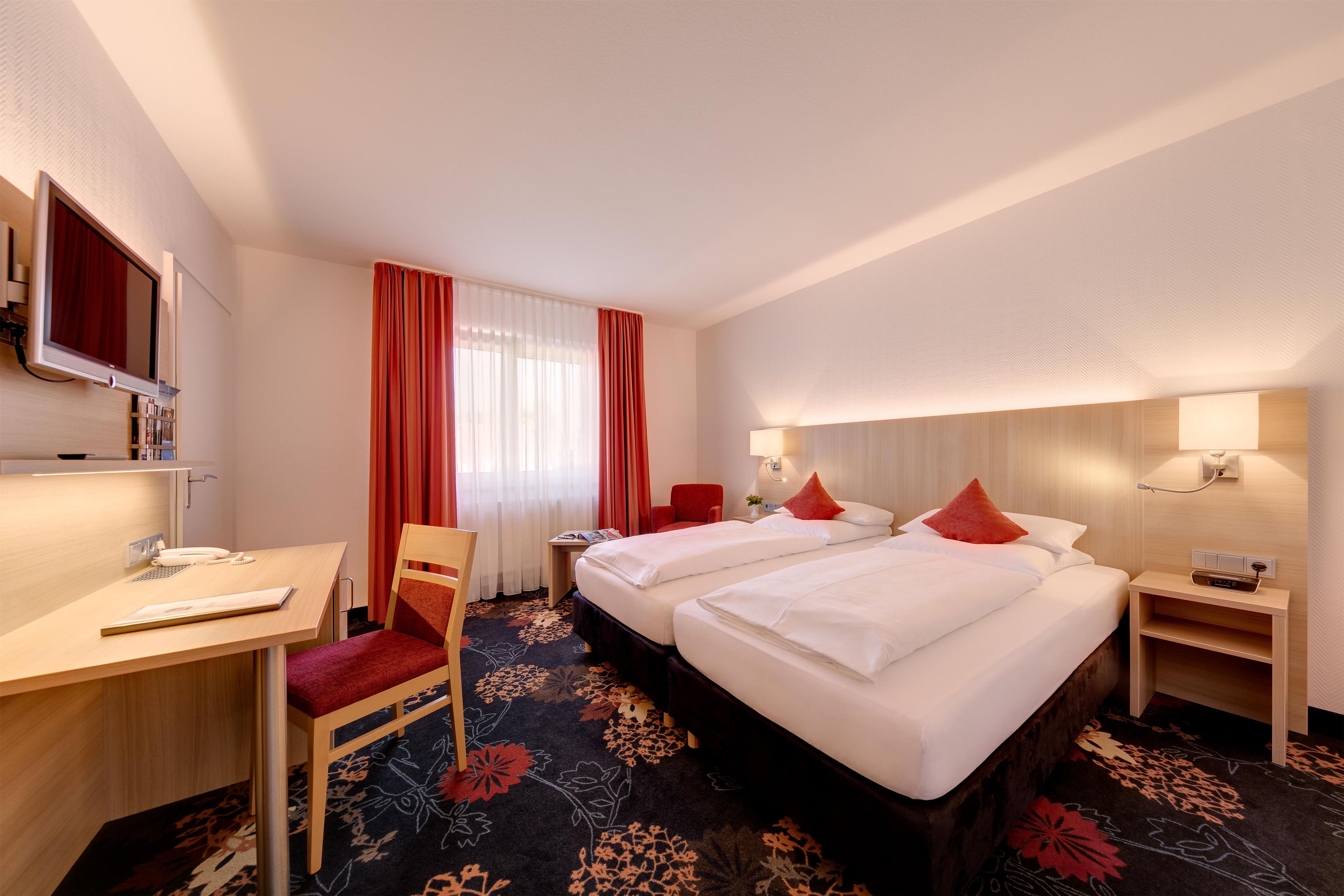 A hotel room with two beds, a desk, a chair, a TV, and a window with curtains.