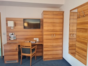 Une pièce de taille modeste, comprenant un bureau en bois, une chaise, un téléviseur fixé au mur et une armoire à deux portes.