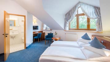 A hotel room with a bed, a desk, a chair, a small table with a vase, and a window with curtains.