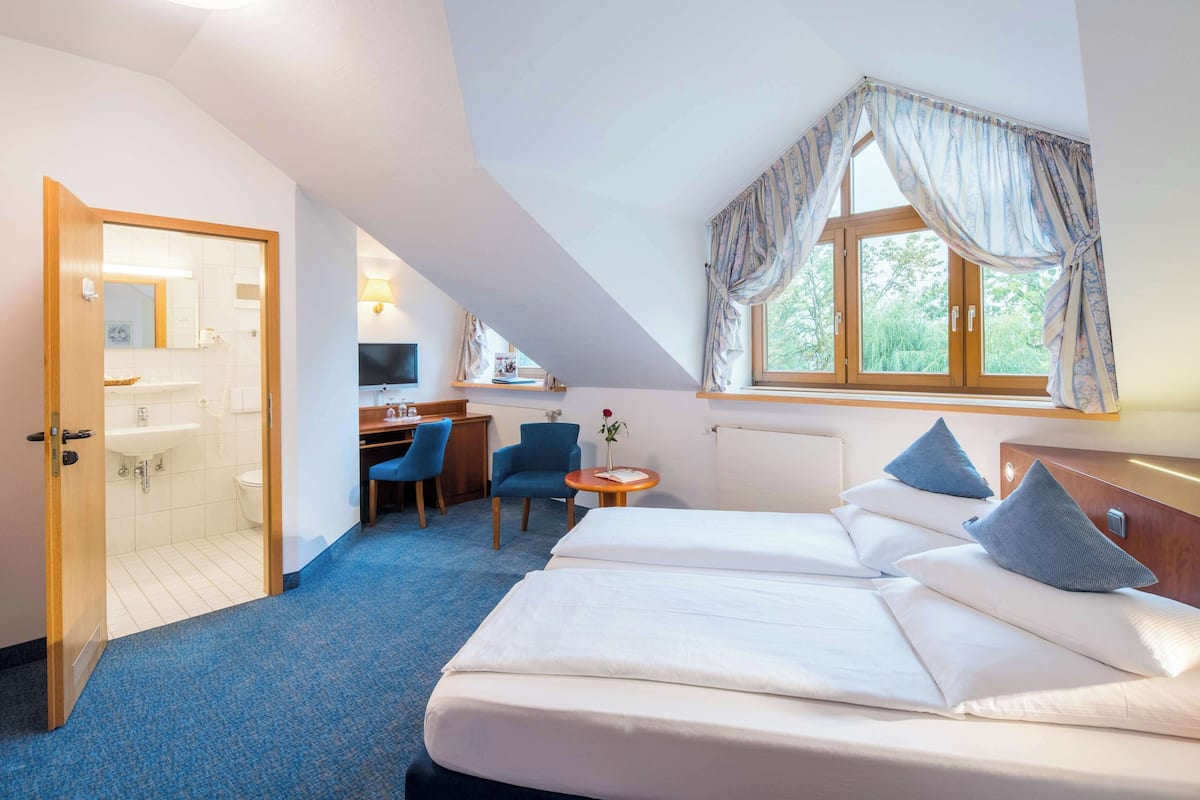 Une chambre d’hôtel avec un grand lit, un bureau, une chaise, une petite table avec un vase et une fenêtre avec des rideaux.