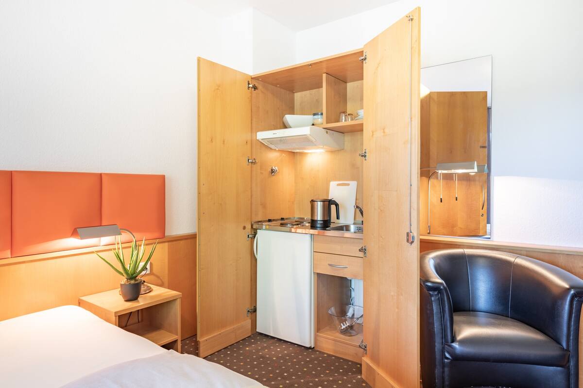 Pieni hotellihuone, jossa on sänky, kirjoituspöytä, jossa on vedenkeitin, tuoli, pieni pöytä, jossa on kasvi, ja peili.