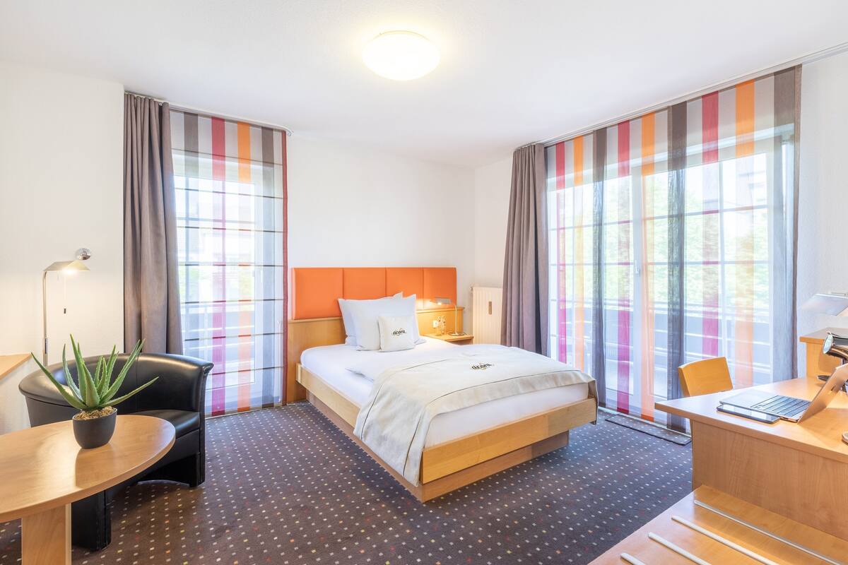 Hotellihuone, jossa on sänky, työpöytä kannettavalla tietokoneella, tuoli, ruukkukasvi ja verhot.