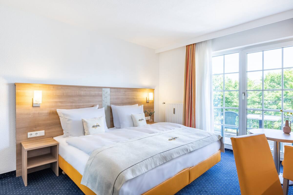 Hotellihuone, jossa on suuri sänky, yöpöytä, pöytä ja tuolit sekä ikkunasta avautuva näkymä vehreään maisemaan.