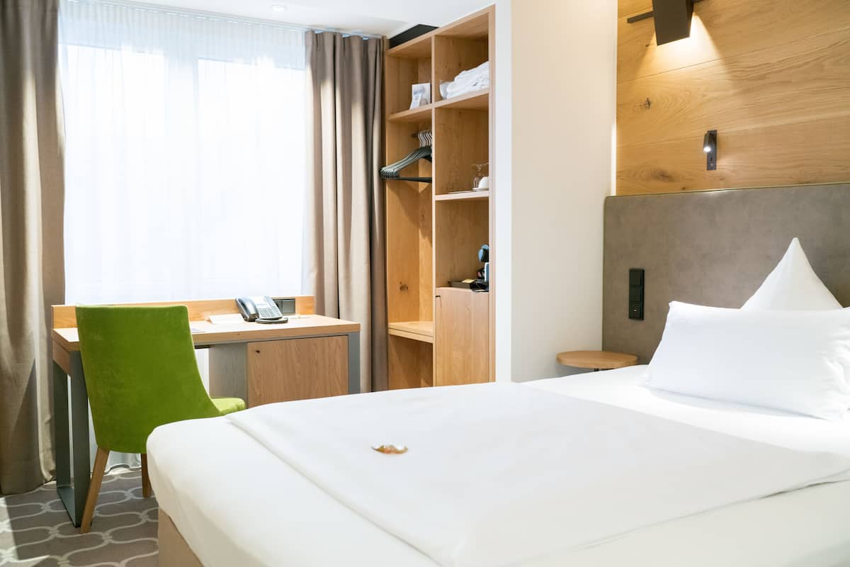 Una habitación de hotel con una cama, un escritorio con teléfono, una silla verde y un armario.