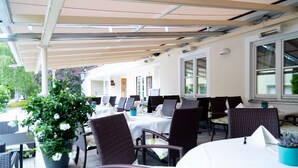 An outdoor seating area with tables and chairs, covered by a canopy, and potted plants.
