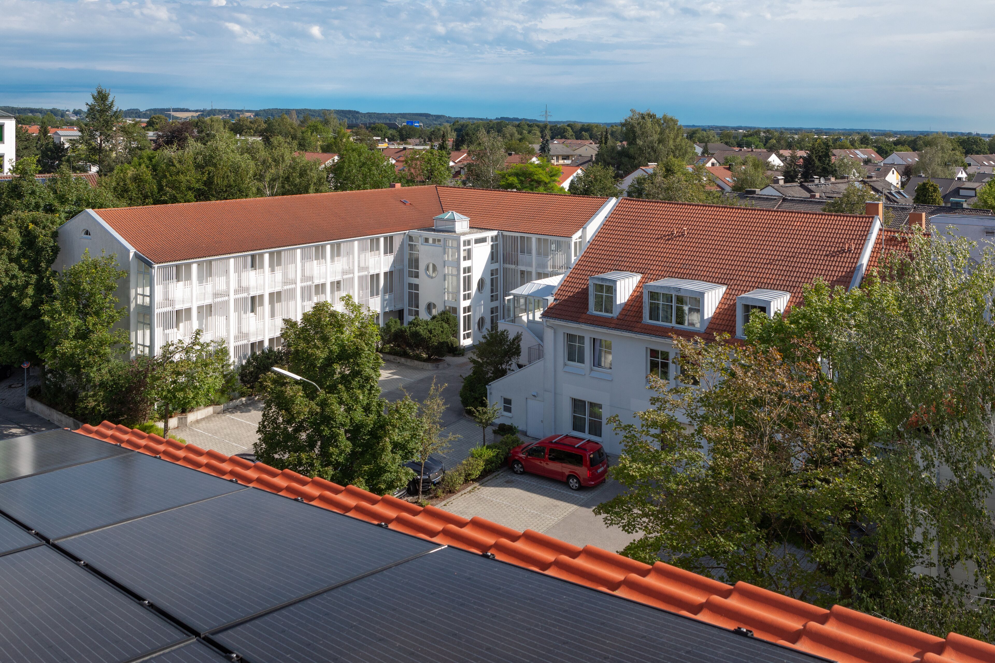 A modern building with a red-tiled roof, surrounded by greenery and other residential buildings.