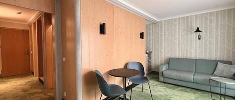 Habitación de hotel con pared decorativa de madera, un sofá verde, una mesa redonda con sillas y una alfombra estampada.