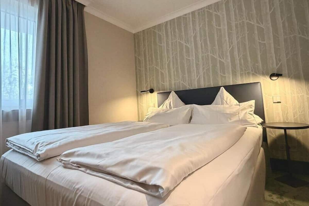 Una cama doble con ropa de cama blanca, cabecera, dos lámparas de noche y una ventana con cortinas.