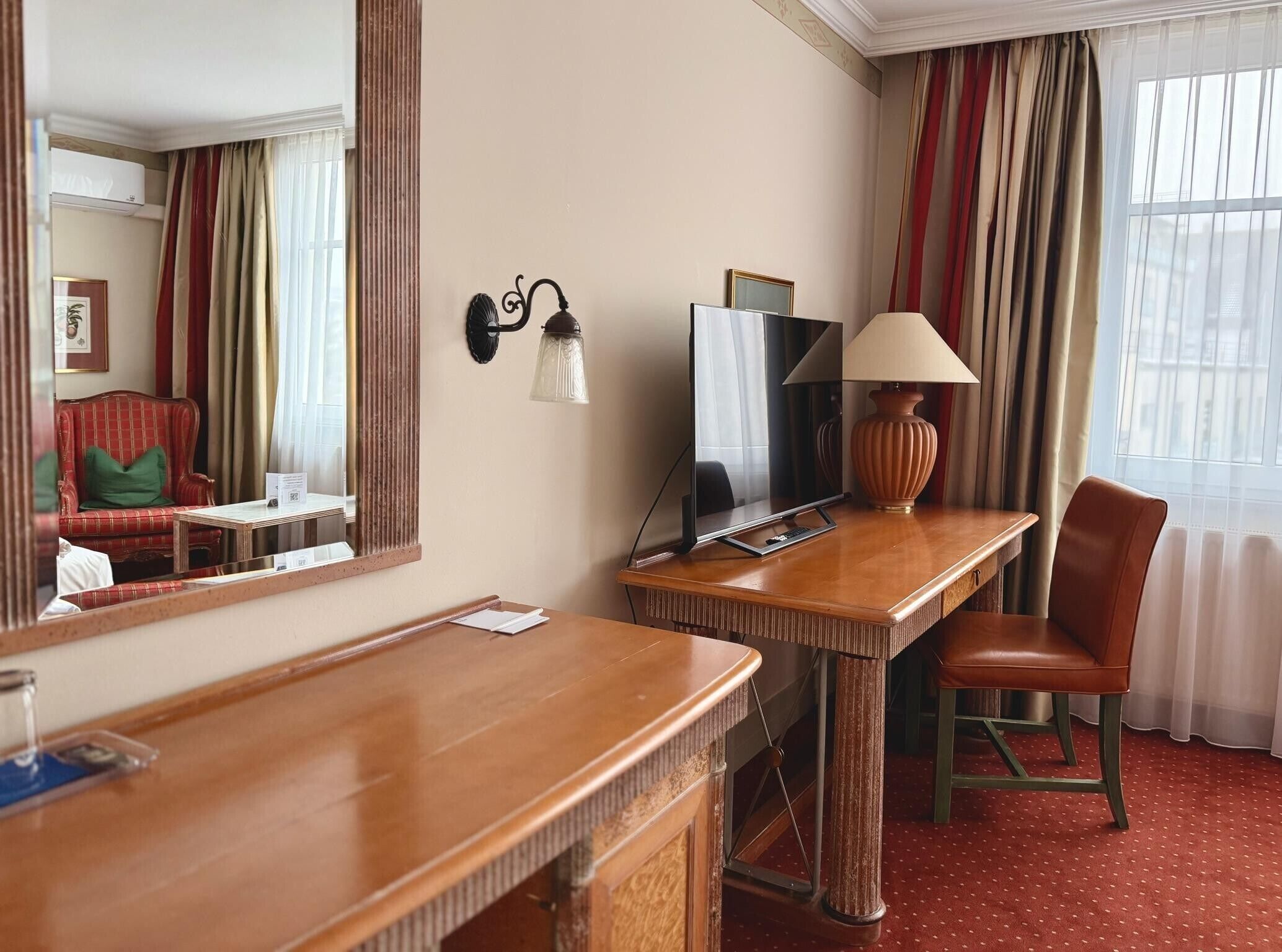 A hotel room with a wooden desk, a chair, a large mirror, a flat-screen TV, a lamp, and a window with curtains.