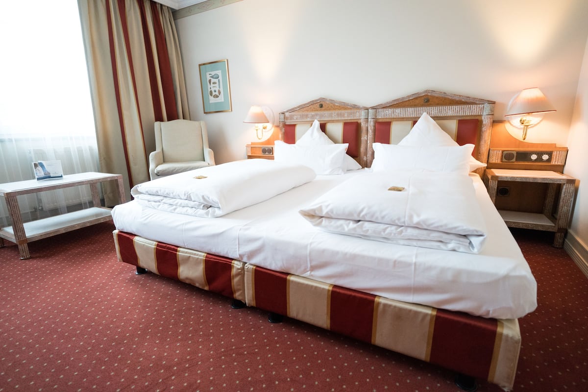 Una cama doble con ropa blanca, un corredor de cama a rayas rojo y beige, dos mesitas de noche con lámparas, una silla y una pequeña mesa con una revista.
