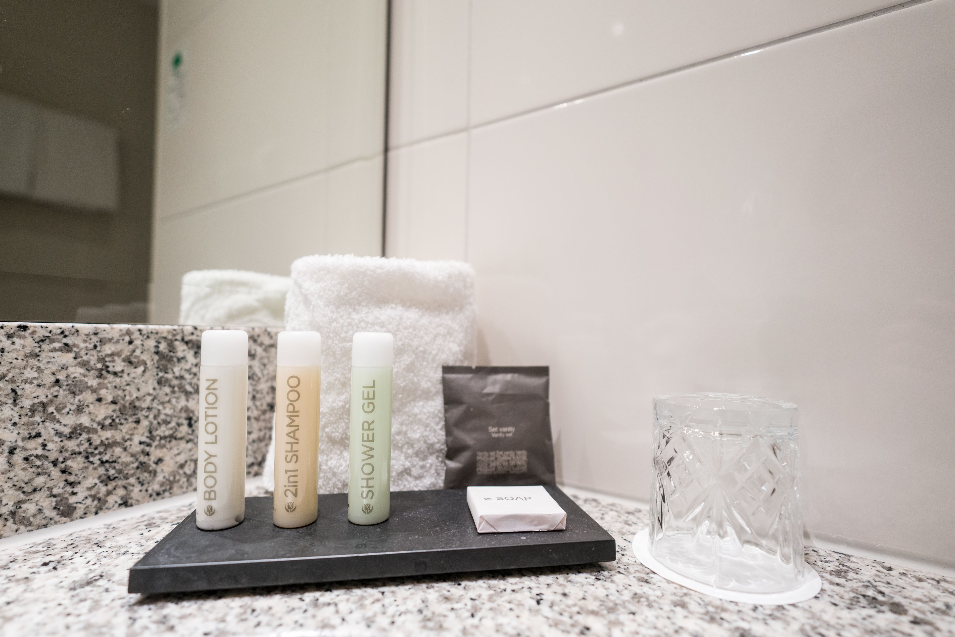 A bathroom counter with toiletries including body lotion, shampoo, and shower gel, a soap bar, and a glass.