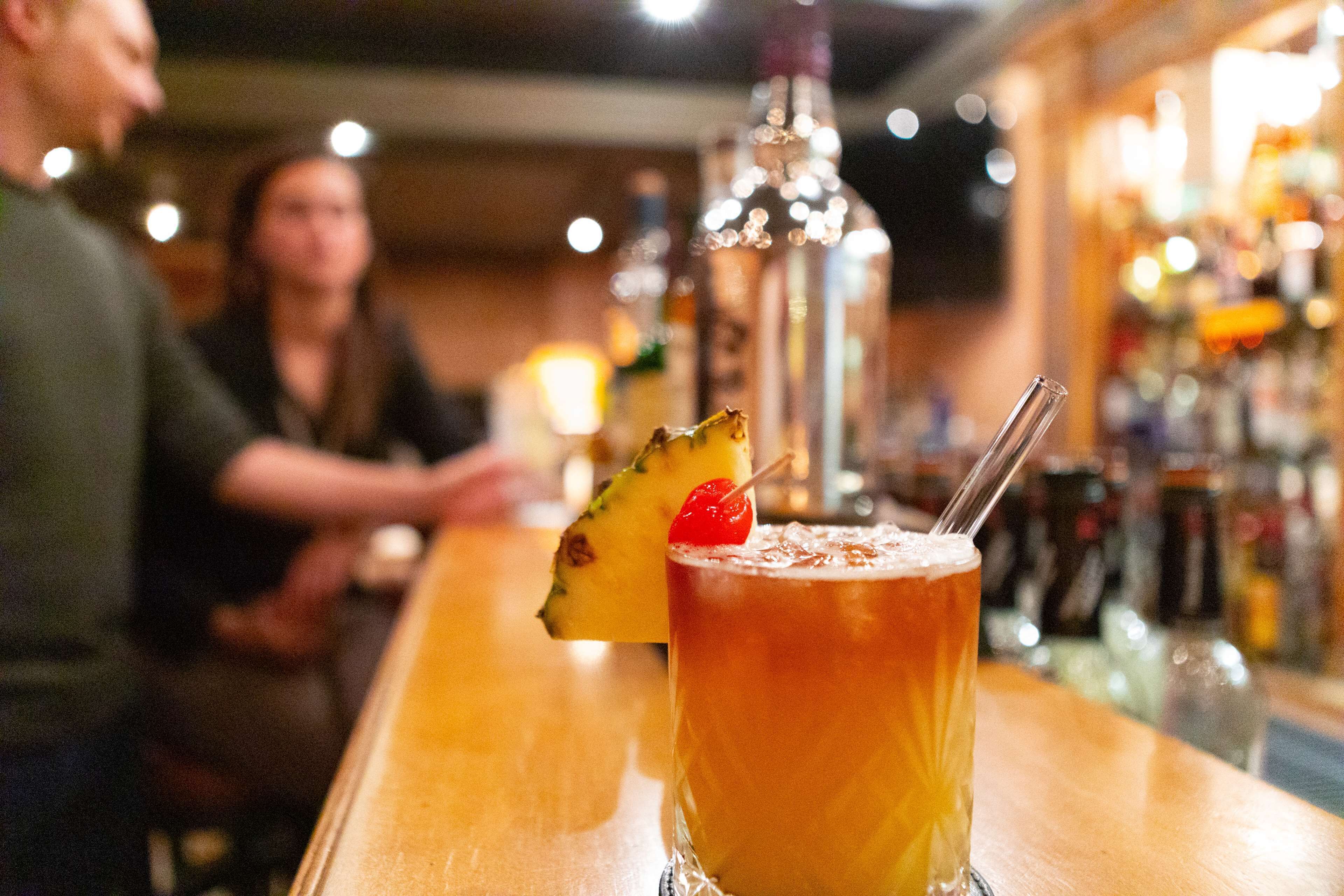 A bar setting with a drink garnished with a pineapple and a cherry, and a bottle of liquor in the background.