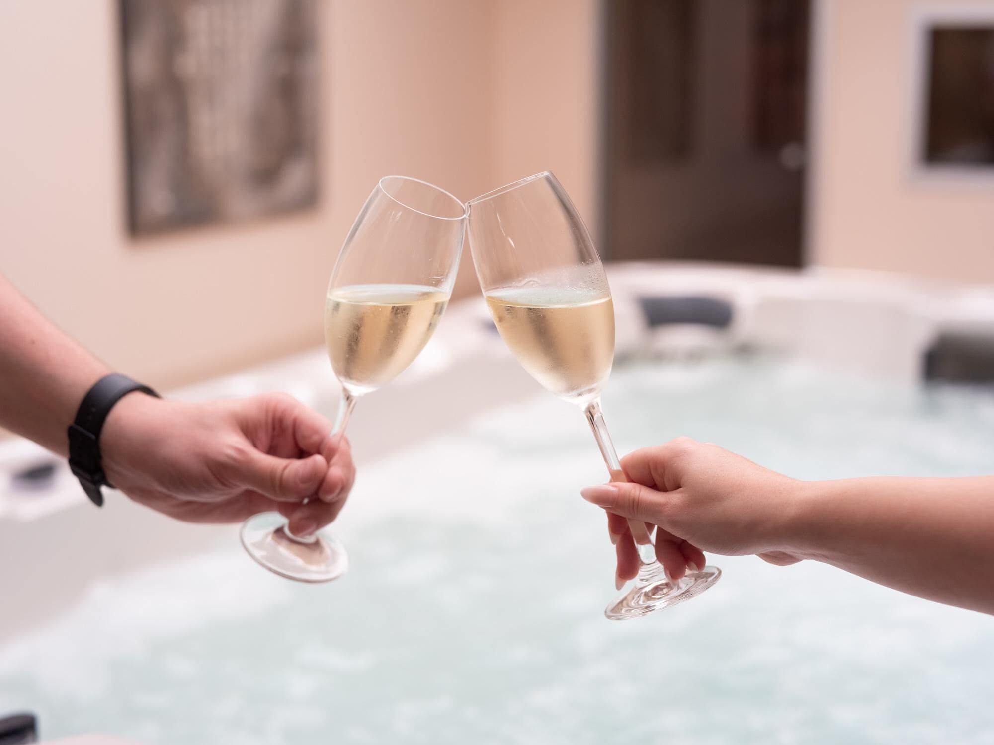 Dos manos sosteniendo copas de vino en una habitación con jacuzzi.