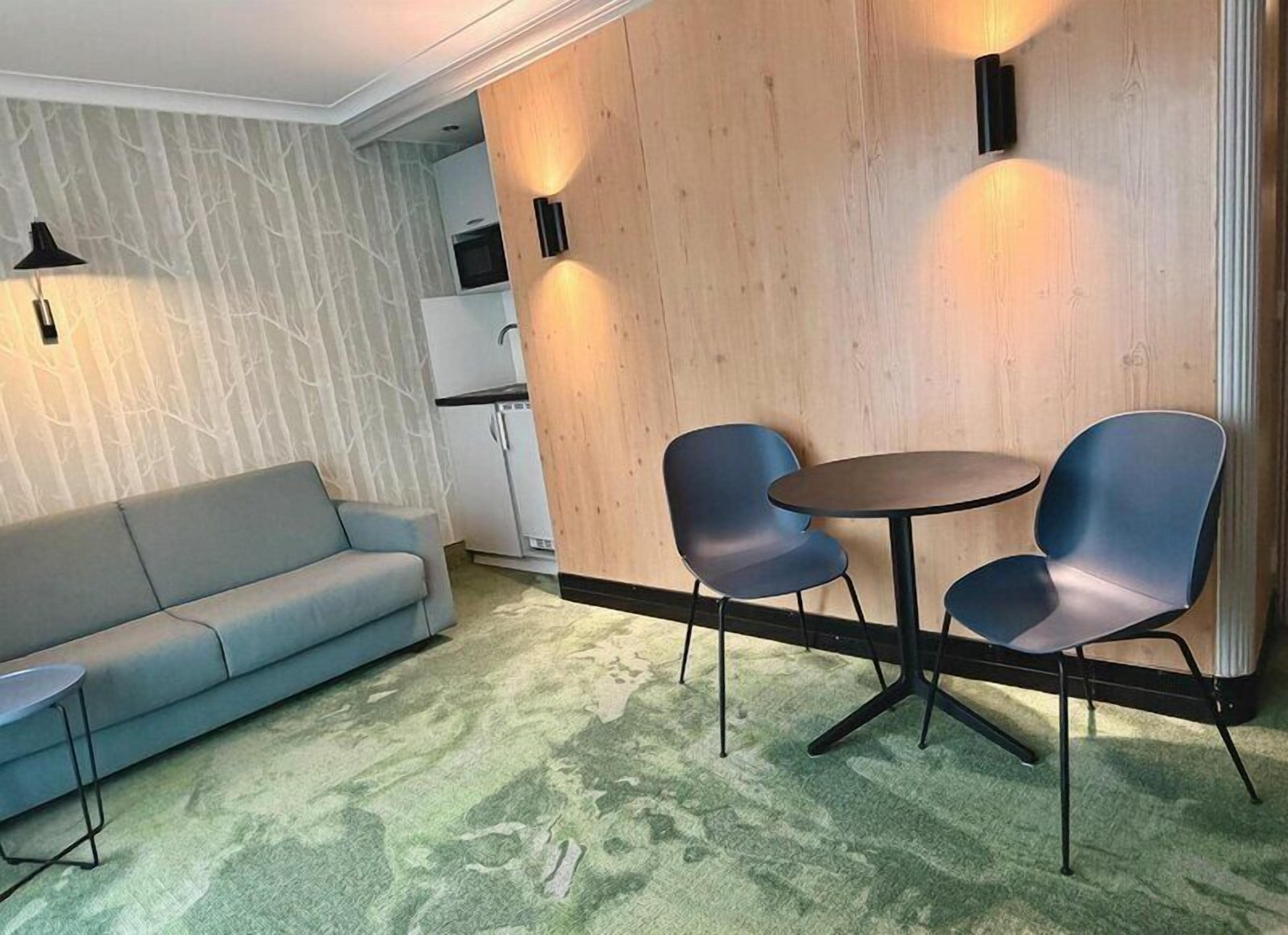 A room with a sofa, two chairs, a small table, a wooden partition, and a kitchen area.