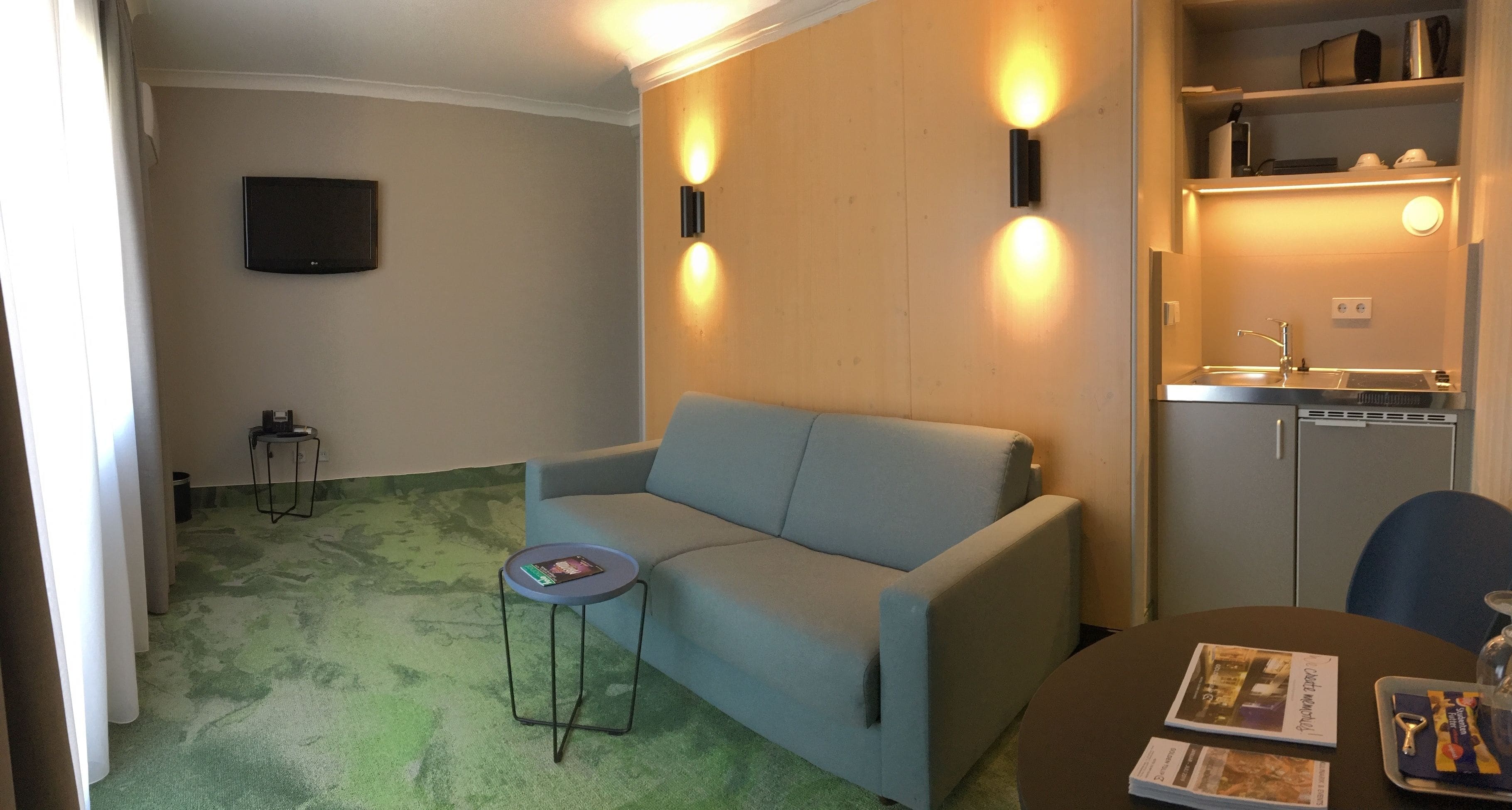 Una sala de estar con un sofá gris, una mesita redonda con revistas, un televisor de pantalla plana montado en la pared y una pequeña zona de kitchenette con un lavaplatos y armarios.