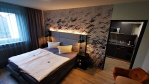Una habitación de hotel moderna con una cama grande, una mesita de noche, una lámpara de noche, suelo de madera, una pequeña zona de estar y una kitchenette.