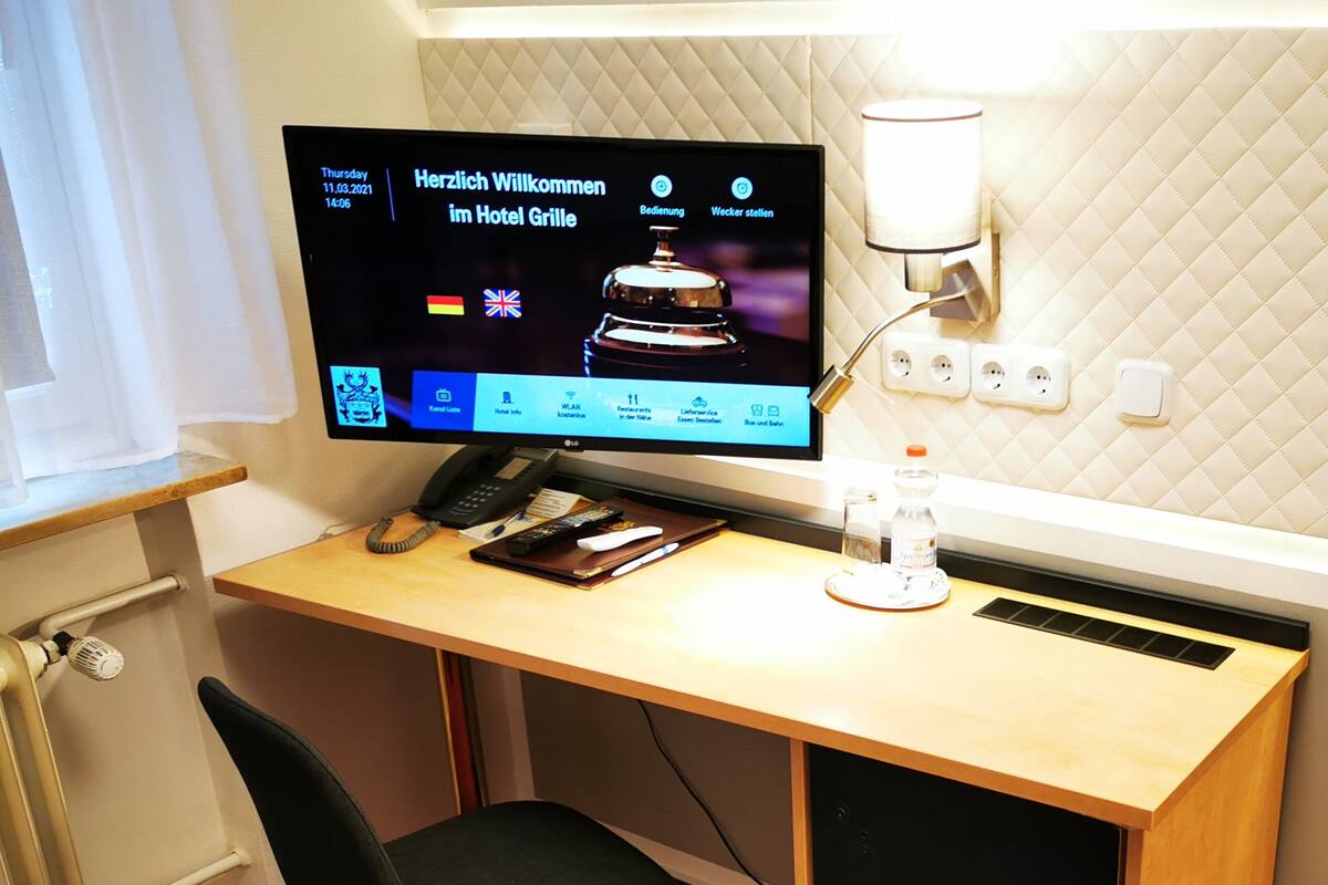 Un bureau dans une chambre d’hôtel, avec un écran d’ordinateur affichant un message d’accueil, un téléphone, une télécommande, une bouteille d’eau et une chaise.